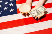 Desde arriba de la cosecha irreconocible persona con bollos mitades y billete de dólar sobre la bandera nacional americana en el Día de la Independencia - foto de stock