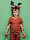 Niño vistiendo cuernos de reno diadema y fiesta festiva ventilador de pie sobre fondo verde y mirando a la cámara - foto de stock