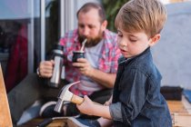 Hipster dad pouring herbal tea from thermos and drinking in calabash gourd against son with hammer working with wood — Stock Photo