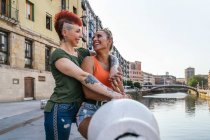 Вид сбоку на веселую молодую гомосексуальную женщину, обнимающую татуированную подружку с ирокезом, глядя друг на друга на канале в городе — стоковое фото