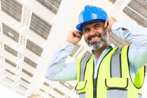 From below of mature happy ethnic male supervisor in vest putting on hardhat while looking away standing near solar power station — Stock Photo