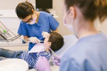 Alto ángulo de dentista y asistente de tratamiento de dientes de niño durante el procedimiento en la clínica de odontología - foto de stock