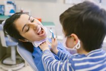 Niño curioso en máscara médica jugando el papel de dentista y la comprobación de los dientes con espejo dental en el hospital - foto de stock