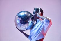 Mujer alegre étnica fresca con pelo corto en auriculares inalámbricos y ropa de moda bailando hip hop con la boca abierta y los ojos cerrados sosteniendo la bola de brillo sobre fondo violeta - foto de stock
