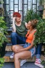Zufriedene junge tätowierte Frau spricht mit homosexuellen Geliebten, während sie sich auf der Treppe zwischen Pflanzen anschaut — Stockfoto
