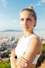 Giovane atleta donna sognante con le braccia incrociate ammirando la città mentre distoglie lo sguardo sotto il cielo blu nuvoloso alla luce del sole — Foto stock