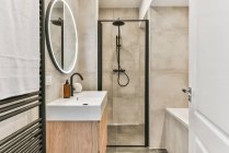 Opened door to tiled bathroom with heated towel rail near shining mirror hanging above sink with soap near stylish shower cabin and bathtub — Stock Photo