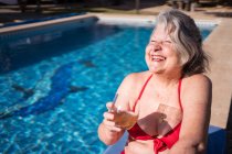 Alto ángulo de mujer turista senior encantada en bikini riendo brillantemente mientras se enfría en la piscina con bebida - foto de stock