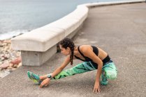 Side view of fit female runner stretching legs and leaning on metal plank while waring up during training on promenade — Stock Photo