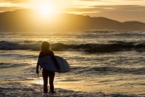 Rückansicht einer unkenntlichen jungen Frau mit Surfbrett, die während des Sonnenuntergangs am Strand in Asturien ins Meer steigt — Stockfoto