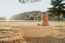 Side view of happy toddler child with toy sitting looking away in plastic bath while playing with water in countryside — Stock Photo
