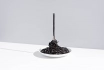 Stainless fork in full bowl of black spaghetti with cuttlefish ink on white table in studio on gray background — Stock Photo