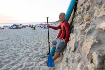 Seitenansicht eines glücklichen männlichen Surfers in Neoprenanzug und Hut, der mit Paddel und SUP-Brett wegschaut, während er sich auf das Surfen an der Küste vorbereitet, angelehnt an eine Steinmauer — Stockfoto