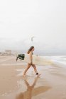 Vista lateral da jovem esportista feliz em roupa de banho com prancha de surf olhando para longe na costa arenosa contra o oceano tempestuoso — Fotografia de Stock