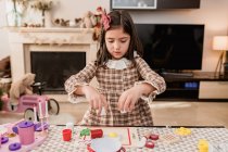 Attentive child in checkered dress playing with toys while breaking egg during cooking process at table in light room — Stock Photo