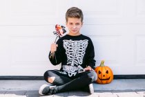 Corpo inteiro de menino pré-adolescente assustador em traje de esqueleto preto com maquiagem segurando flor e olhando para a câmera enquanto sentado perto de lanterna de abóbora contra a parede branca — Fotografia de Stock