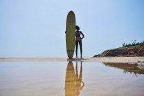 Visão traseira da atleta afro-americana com prancha de surf admirando o oceano da costa arenosa sob o céu azul nublado — Fotografia de Stock
