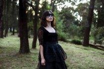 Мистическая ведьма в длинном черном платье с раскрашенным лицом, смотрящая в темные мрачные леса — стоковое фото