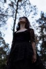 From below mystic witch in long black dress and with painted face standing looking away in dark gloomy woods — Stock Photo