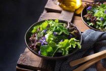 De arriba de la ensalada sabrosa vegetariana con las hojas verdes y rojas de lechuga y las flores comestibles contra la jarra del aceite - foto de stock