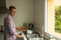 Vista lateral de la calma reflexiva madura lavando platos sucios mientras está de pie cerca del fregadero en la cocina y haciendo las tareas domésticas - foto de stock