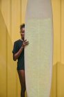 Jovem desportista afro-americana alegre de biquíni e t-shirt olhando para a câmera com prancha de surf em um recipiente amarelo na costa — Fotografia de Stock