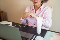Conteúdo da colheita empresária idosa em fones de ouvido aplicando creme hidratante na mão enquanto tem videochamada no laptop no escritório — Fotografia de Stock