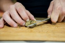 Cultivo masculino irreconocible con cuchillo que corta la pieza seca de la planta de cannabis en el tablero de madera en el espacio de trabajo - foto de stock
