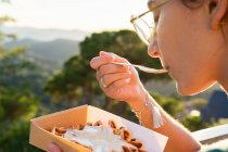Вид сбоку на урожай молодая женщина ест вкусные бельгийские вафли со взбитыми сливками в коробке для еды на вынос против монтировок в задней освещении — стоковое фото