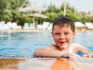 Deleitado niño lindo con el pelo mojado apoyado en la piscina y mirando a la cámara mientras se divierten durante el fin de semana de verano - foto de stock