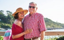 Seniorenpaar steht dicht beieinander und blickt in die Kamera und genießt den Blick auf das Meer an einem sonnigen Tag — Stockfoto