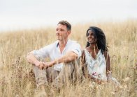 Homme souriant adulte avec une femme ethnique bien-aimée se reposant sur le pré tout en passant le week-end à la campagne — Photo de stock
