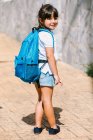 Rückenansicht eines Schulkindes mit Rucksack auf dem Bürgersteig, das im Sonnenlicht der Kamera über die Schulter schaut — Stockfoto
