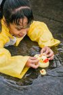 Чарівна етнічна дитина в плащі грає з пластиковими качками, що відображаються в рваній калюжі в дощову погоду — стокове фото
