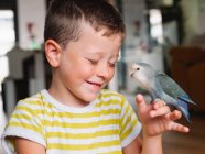 Симпатичный маленький мальчик в полосатой футболке сидит с маленькой птичкой с серым оперением дома — стоковое фото