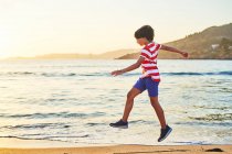 Visão lateral comprimento total do menino correndo na costa arenosa lavado por mar espumoso — Fotografia de Stock