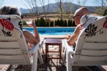 Visão traseira do casal idoso irreconhecível em trajes de banho beber café gelado enquanto relaxa em espreguiçadeiras no quintal — Fotografia de Stock