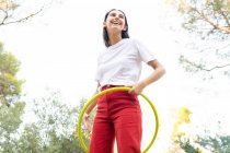 Par en bas de joyeux adolescent féminin en jean rouge tournoyant hula hoop tout en ayant du temps libre dans le parc — Photo de stock