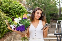 Zufriedene junge Frau in Brille mit blühendem Blumenstrauß SMS-Nachrichten auf dem Handy auf städtischen Treppen — Stockfoto