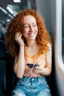 Mulher jovem cândida com cabelo encaracolado vermelho e celular ouvindo música de fones de ouvido enquanto olha para longe no trem — Fotografia de Stock