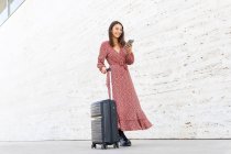 Positive female in long red dress walking with luggage on street against white wall in daytime — Stock Photo