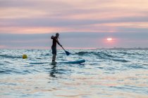 Vista lateral do surfista masculino de fato de mergulho e chapéu na prancha de remo surfando na praia durante o pôr do sol — Fotografia de Stock