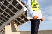 Low angle of cropped unrecognizable mature ethnic male inspector in vest with clipboard looking away while standing near solar power station — Stock Photo
