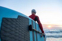 Vue arrière d'un surfeur masculin méconnaissable en combinaison et chapeau portant une planche à pagaie et entrant dans l'eau pour surfer sur le bord de la mer — Photo de stock