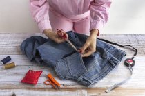 Crop anonymous female seamstress using scissors while cutting jeans pocket on wooden table in atelier — Stock Photo