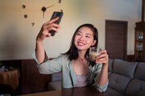 Zufriedene junge ethnische Frau beim Selbstporträt mit Kaffee auf dem Handy, während sie zu Hause auf dem Sofa mit Laptop sitzt — Stockfoto
