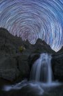 Viajante admirando cascata com espuma em montagem áspera contra lagoa sob céu estrelado em movimento ao entardecer — Fotografia de Stock
