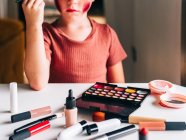 Cropped unrecognizable charming child with makeup applicator touching head while looking away at table with eyeshadow palette — Stock Photo