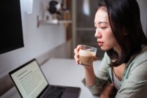 Вид сбоку молодой этнической блоггерши за столом с нетбуком и кофе, смотрящей на камеру в комнате дома — стоковое фото
