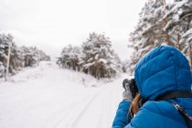 Jovem fêmea em outerwear quente em pé entre árvores nevadas e tirar foto na câmera na floresta de inverno — Fotografia de Stock